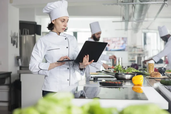 拥有现代笔记本电脑的高级厨师站在餐厅的专业厨房里寻找美食菜谱. — 图库照片