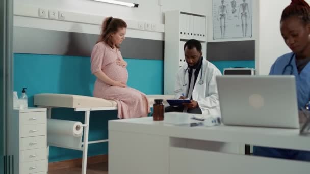 Médico afroamericano tomando notas en el examen con la futura madre — Vídeos de Stock
