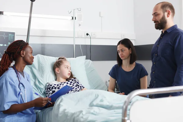 Bezorgde jonge ouders luisteren naar Afrikaans-Amerikaanse verpleegster praten over overleg resultaten van ziek klein meisje. — Stockfoto