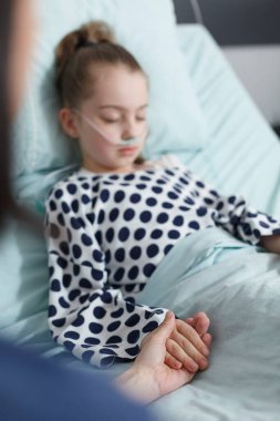 Bakıcı anne hasta kızının elini tutarken çocuk kliniği yatağında dinleniyor.