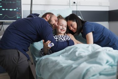 Pediyatri hastanesindeki hasta odasında iyileşmekte olan küçük kızı kucaklayan mutlu genç ebeveynler..