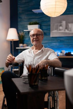 Gülümseyen yaşlı gri saçlı adamın portresi. Masanın önünde elinde kalem tutarak kendine güvenen bir poz veriyor.