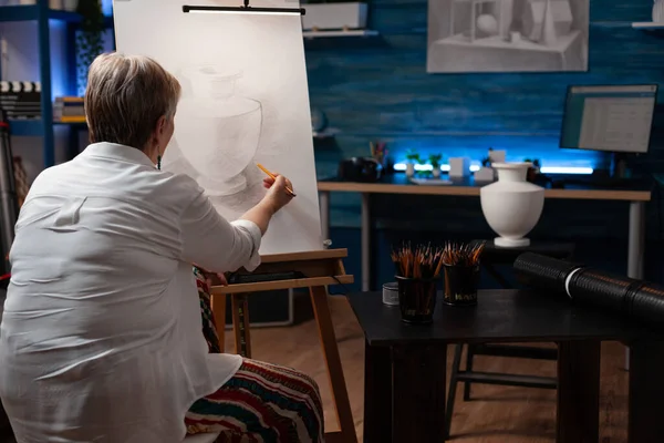 Starsza kobieta czuje inspirację trzymając ołówek rysunek wazon model na papierze na sztaludze — Zdjęcie stockowe