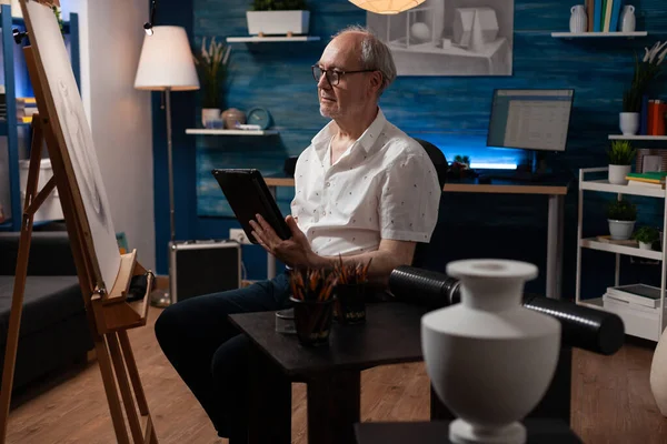 Emekli sanatçı dijital tablete göz atıyor kalem çizimiyle resim sehpasına bakıyor ve ilham buluyor.