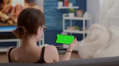 Elinde yeşil ekranlı yatay akıllı telefon tutan genç bir kadın video içeriği izleyen bir uygulama kullanıyor.