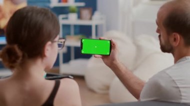 Yakından, elinde yeşil ekranlı akıllı telefon olan bir adamın kız arkadaşıyla çevrimiçi video içeriği izlediği.