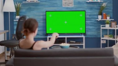 Kanepede oturan genç kadın TV 'de yeşil ekrana bakıyor ve kanal değiştiriyor.