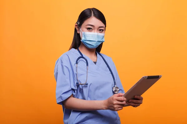 Clinica sanitaria infermiera asiatica indossa maschera antivirale e stetoscopio pur avendo tablet moderno. — Foto Stock