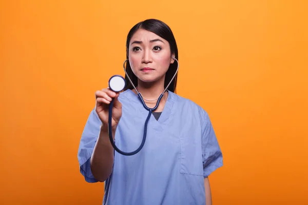 Dotknięta azjatycka pielęgniarka używająca stetoskopu do konsultacji z chorym pacjentem na pomarańczowym tle. — Zdjęcie stockowe