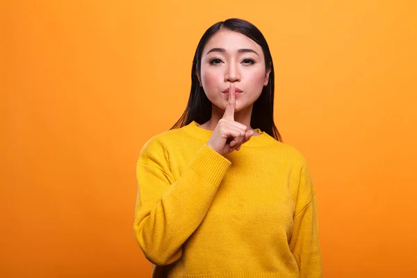 Beautiful mysterious woman wearing vibrant yellow sweater making silence gesture on orange background. — 스톡 사진