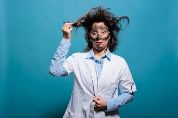 Wacky and crazy looking mad scientist grabbing messy hair while having dirty face from explosion on blue background. — стоковое фото