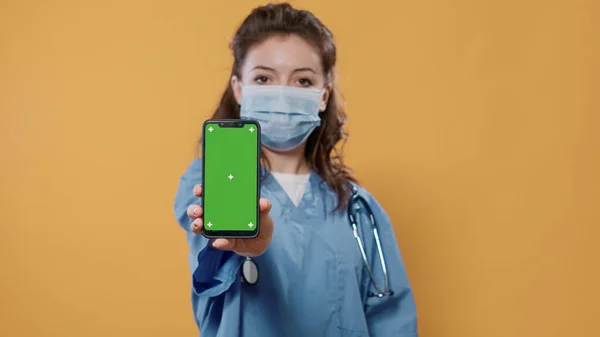 Portrait of medic in hospital uniform wearing surgical mask holding smartphone and texting showing green screen mockup — 스톡 사진