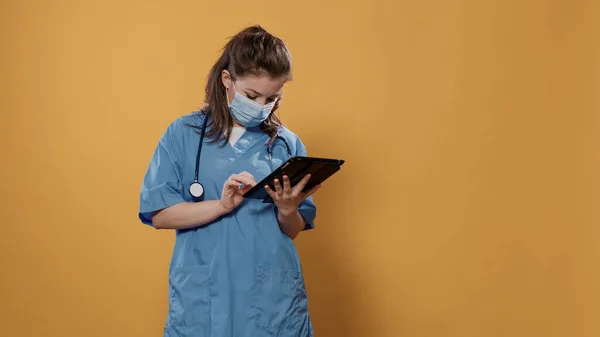 Portrait of woman doctor holding tablet typing patient data wearing hospital uniform and covid protection mask — 스톡 사진