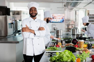 Lokantanın profesyonel mutfağında kameraya gülümserken kolları çapraz duran gastronomi uzmanı..