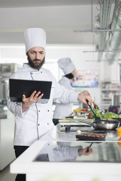 Chef preparing fresh organic vegetable garnish using pan and spatula while looking at laptop screen.
