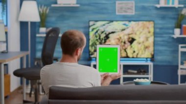 Man sitting on sofa holding vertical digital tablet with green screen watching online social media content