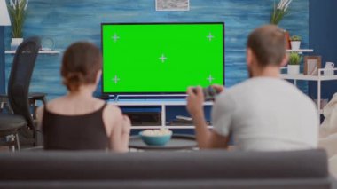 Couple holding controllers playing console online games on green screen tv sitting on couch