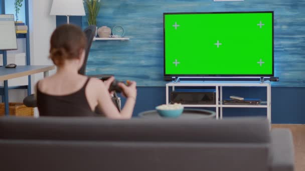 Static tripod shot of gamer girl holding wireless gamepad playing action console video game on green screen tv — Video