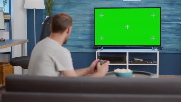 Человек, держащий беспроводной контроллер, играющий в консольную видеоигру на зеленом экране телевизора, сидя на диване — стоковое видео
