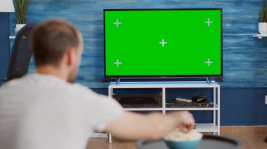 Yeşil ekran TV 'de maç izleyen taraftarların omuz manzarası kanepede oturan favori takımı cesaretlendiriyor. Erkek sporcu oturma odasında krom anahtar ekranlı televizyona bakıyor.