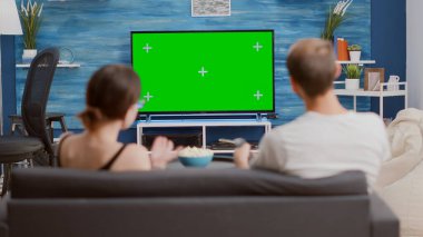 Çift kanepede oturup televizyon izler yeşil ekran düz ekran TV 'de favori programdan ve modern oturma odasında zaplamaktan bahseder. Krom anahtar ekranında patlamış mısır yiyen erkek ve kadın..