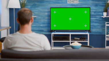 Kanepede oturan bir adam, yeşil ekrana bakıyor, televizyon izliyor, modern oturma odasında bir kase patlamış mısırla film seyrediyor. Krom anahtarla televizyonun önünde oturan kişinin arka planı.