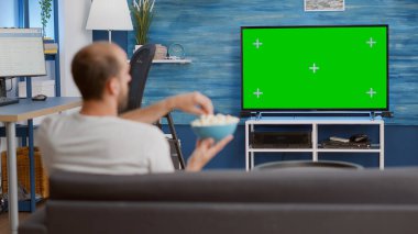 Yeşil ekranda en sevdiği TV programını izlerken kaseden patlamış mısır yiyen adam koltukta rahat oturuyordu. Kanepede oturan kişi oturma odasındaki kroma anahtar ekranında televizyon programını izliyor.