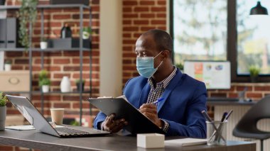 Şirket çalışanı, iş projesi yaratmak için çevrimiçi belgelerle ilgili rapor yazıyor. Yüz maskeli bir ofis çalışanı, dizüstü bilgisayardaki istatistik verilerini planlamak ve interneti taramak için evrak işleri hakkında notlar alıyor..