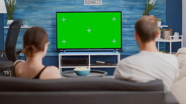 Static tripod shot of couple sitting on sofa looking at green screen on tv relaxing watching movie with bowl of popcorn. Back view of man and woman sitting on couch in front chroma key display.