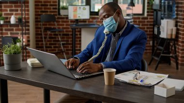 Young businessman with face mask talking to people on landline phone, answering call from project manager. Chatting on office telephone for remote conversation, dialing number line.