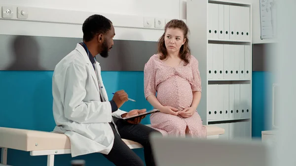 Male physician having conversation with future mother in cabinet, taking notes on papers to give prescription medicine. General practitioner giving pregnancy advice to woman expecting child.
