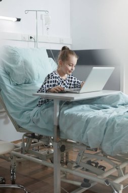 Hasta küçük çocuk hastane yatağında dinlenirken dizüstü bilgisayar oyunlarından zevk alıyor..