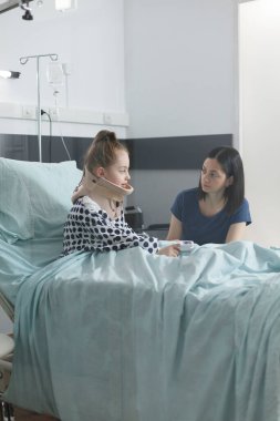 Stresli ebeveyn, pediatri kliniği tedavi odasında otururken yaralı küçük kızıyla konuşuyor..