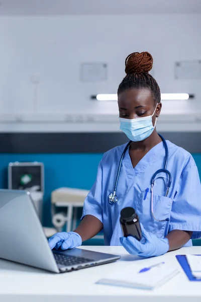 Wearing facemask nurse using modern computer to read prescribed antibiotic prospectus — Stock Photo, Image