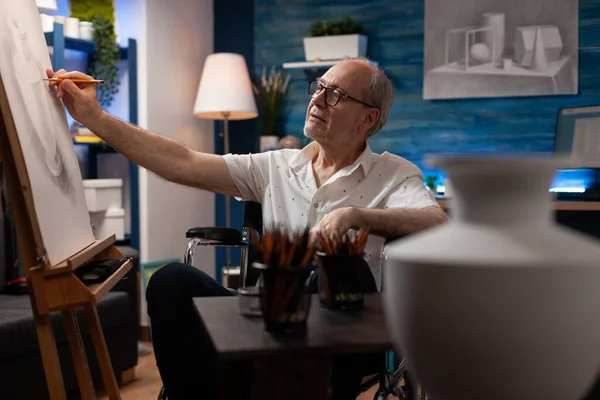 Ανώτερος άνθρωπος συνταξιούχος καλλιτέχνης χρησιμοποιώντας αναπηρική καρέκλα κάνει σκίτσο μολύβι του μοντέλου βάζο μπροστά από το τραπέζι με μολύβια — Φωτογραφία Αρχείου
