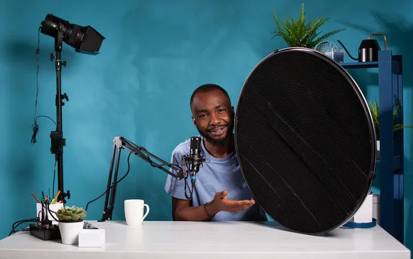 Vlogger explaining features of studio flash light modifier sitting at desk with microphone — Foto de Stock