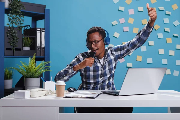 Animado trabalhador de escritório afro-americano alegre usando fones de ouvido sem fio enquanto faz karaoke sentado na mesa. — Fotografia de Stock