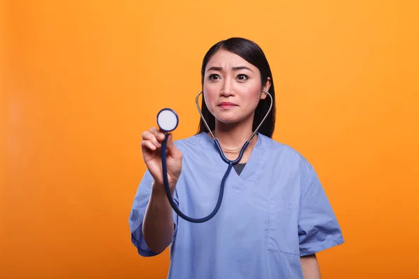 Inquiète asiatique soignant utilisant stéthoscope pour consulter le patient sur fond orange. — Photo