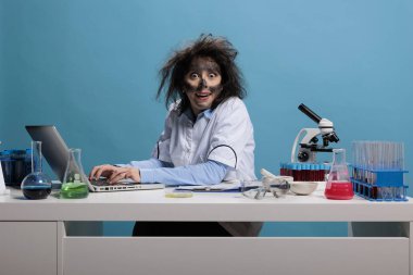 Çılgın aptal kimyager masasında oturmuş laptopta çalışırken mavi arka plandaki kameraya sırıtıyor..