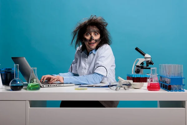 Crazy silly chemist with wild look sitting at desk working on laptop while grinning creepy at camera on blue background. — 스톡 사진