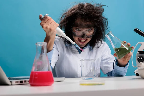 Crazy mad scientist mixing liquid substances while grinning dreadful and sitting at desk on blue background.