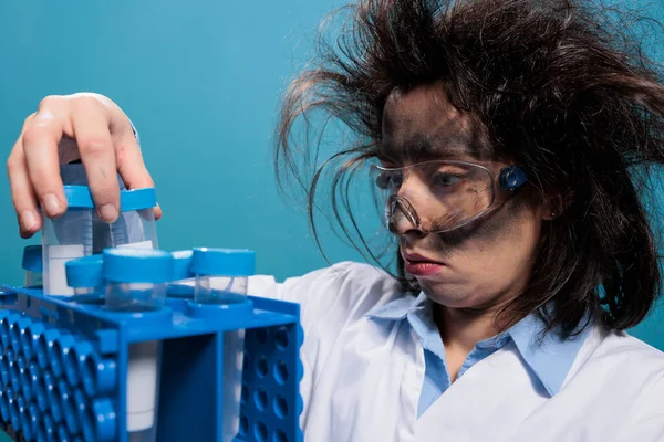 Crazy scientist with dirty face and messy hair analyzing plastic test flasks after laboratory chemical explosion.