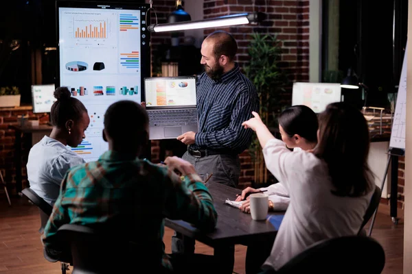 Работник офиса держит ноутбук с бизнес-презентацией, глядя на экран, в то время как коллега указывает на диаграммы — стоковое фото