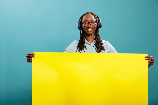 Kablosuz modern kulaklıkları varken elinde sarı kağıt pankart tutan mutlu kadın. — Stok fotoğraf