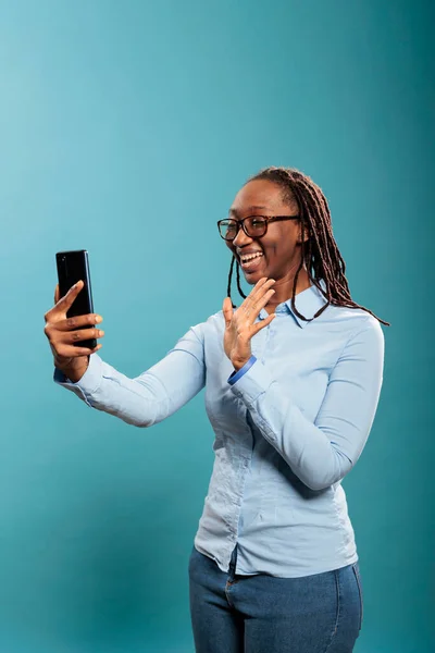 Beautiful confident young lady greeting friend over online phone video call while standing on blue background. — Stockfoto