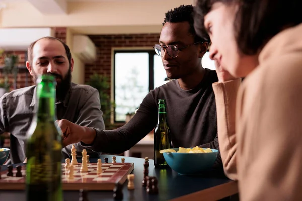 Homem de aparência inteligente movendo peça de xadrez a bordo enquanto sentado à mesa na sala de estar. — Fotografia de Stock