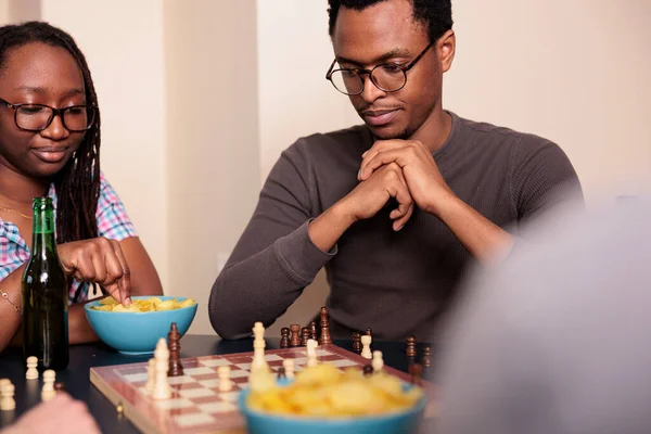 Pensive friends sitting at table in living room while thinking about next chessboard game move. — Stock Photo, Image