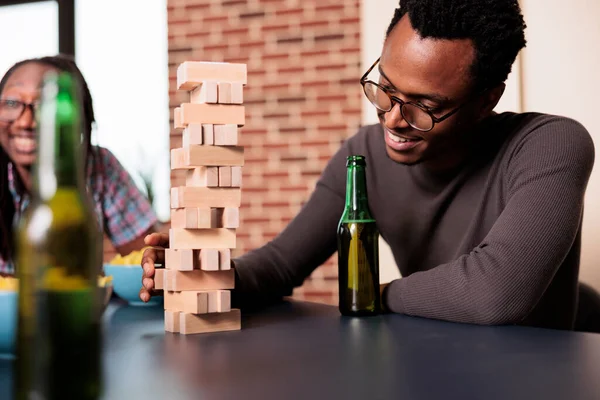 Happy man with beverage on table carefully removing wooden block from wood tower. — Φωτογραφία Αρχείου