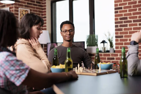 Homem sorridente sentado à mesa na sala de estar com os amigos enquanto assiste a um jogo de xadrez. — Fotografia de Stock