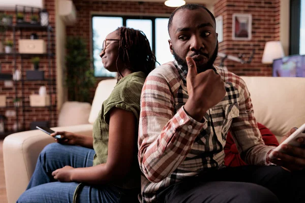 Casal afro-americano perturbado segurando smartphones discutindo sobre viver juntos sentados no sofá — Fotografia de Stock
